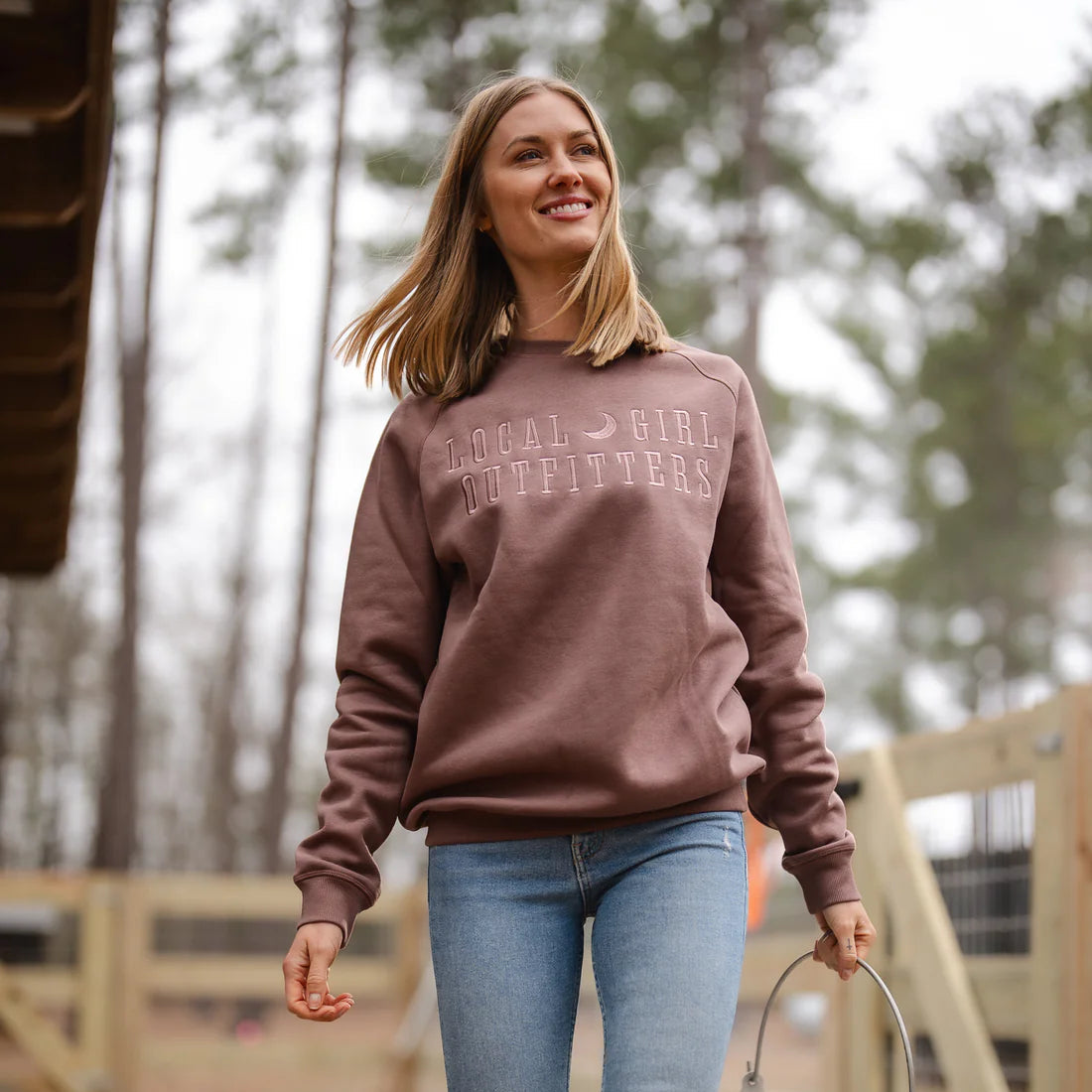Woman in a brown sweatshirt and jeans holding a metal bucket in a forested area.