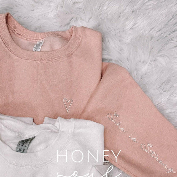 Two sweatshirts, one pink and one white, on a fluffy white surface with text on them.