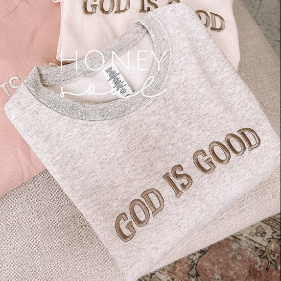 Three folded sweatshirts with 'GOD IS GOOD' text on a textured surface.