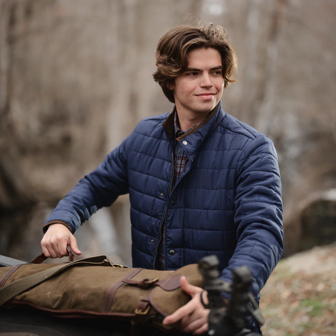 Man in a blue jacket holding a bag outdoors with trees in the background