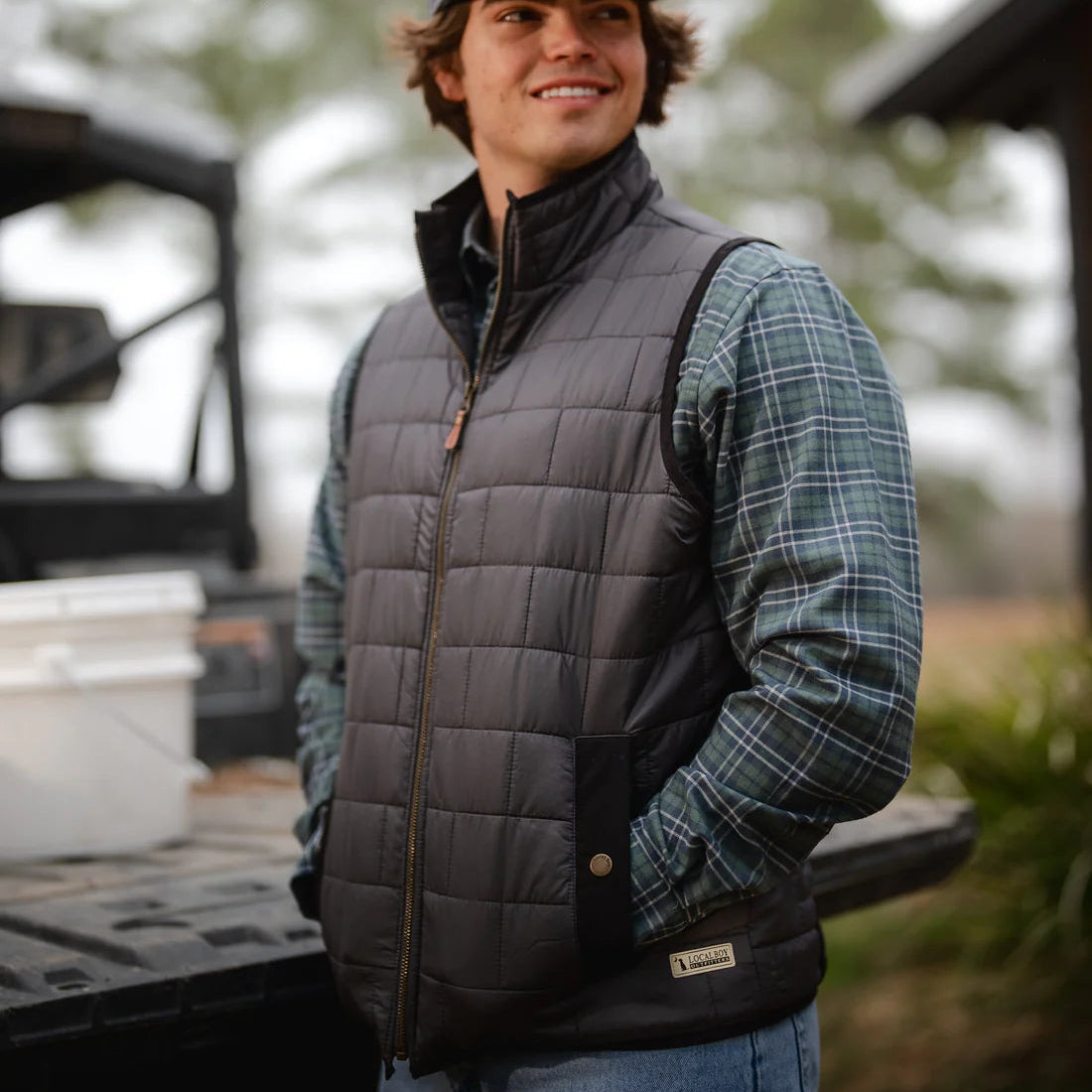 Person wearing a gray quilted vest and plaid shirt outdoors with trees in the background