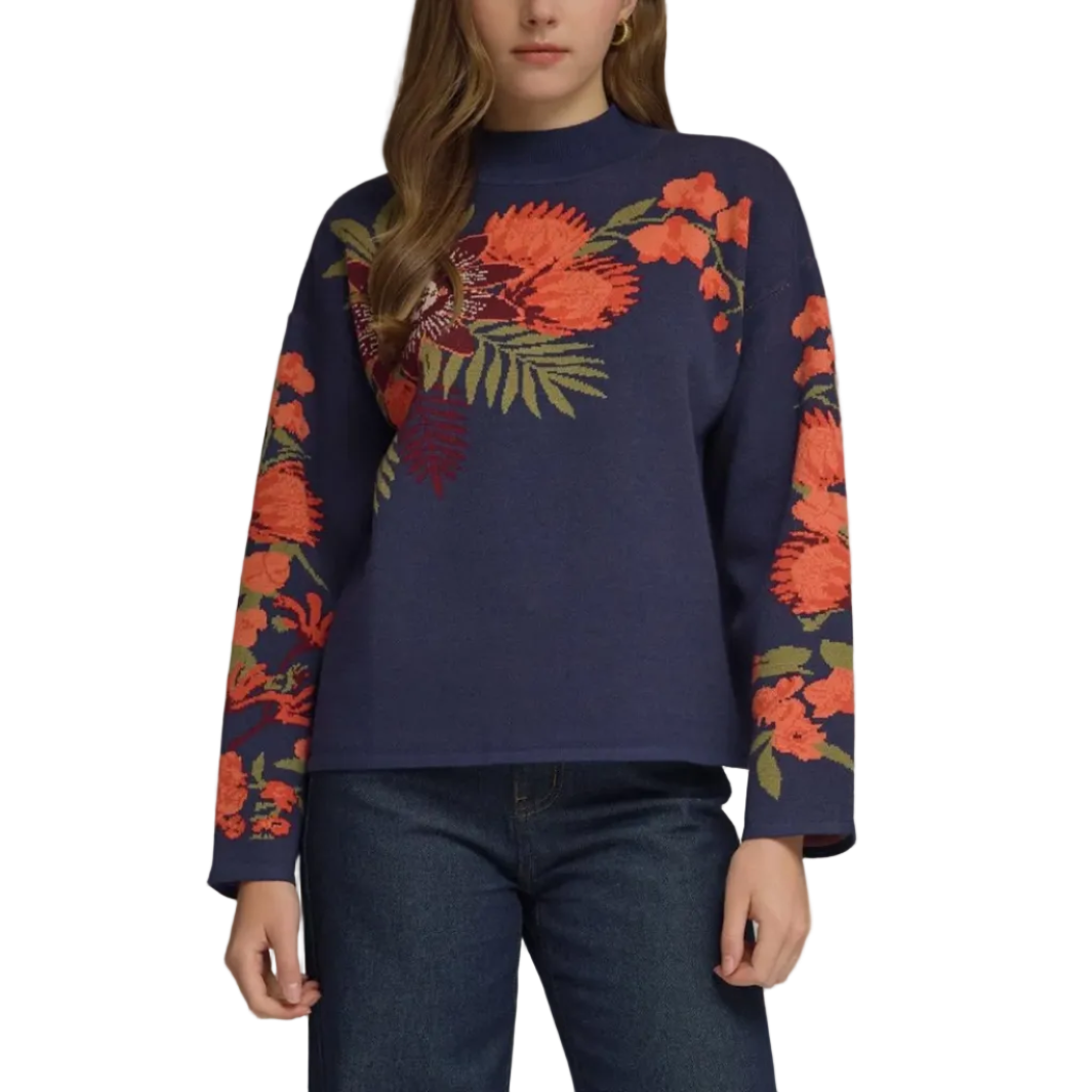 A woman modeling a navy blue crewneck sweater with vibrant orange and red floral patterns on the chest and sleeves, paired with dark blue jeans, set against a plain gray studio background.