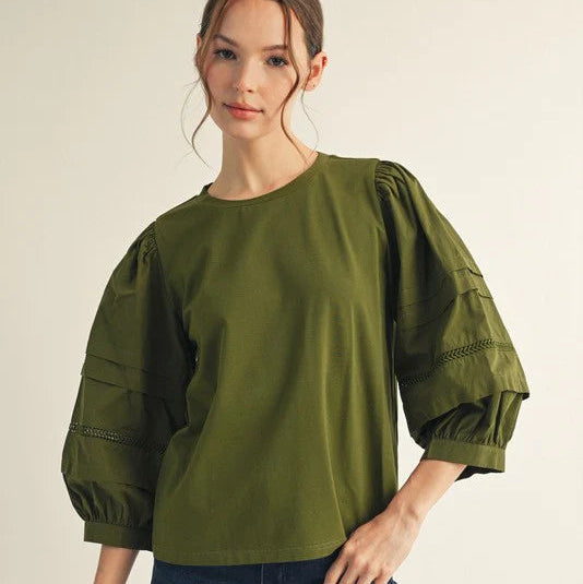 Woman wearing an olive green blouse with puffed sleeves and blue jeans on a beige background